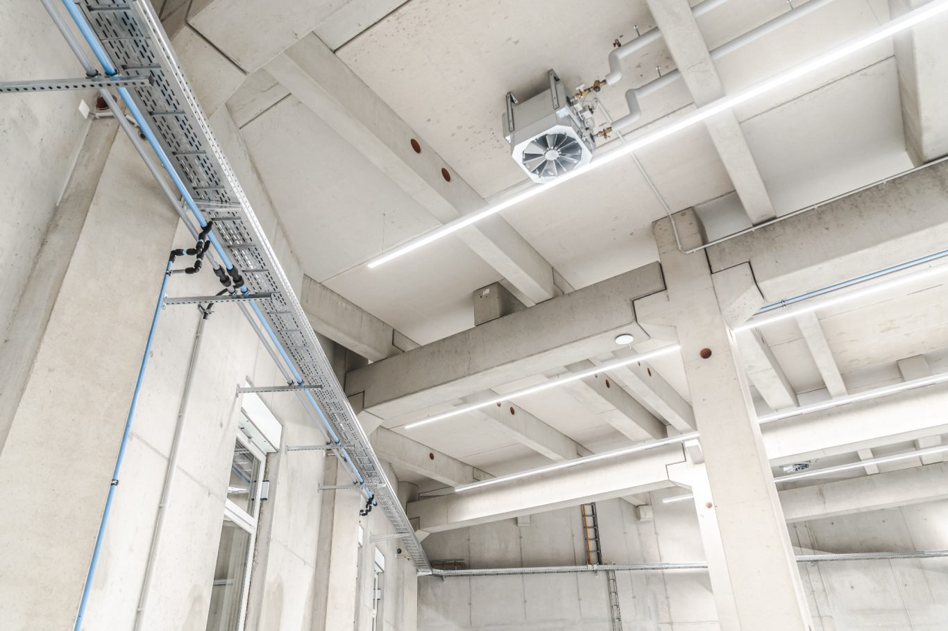 Hallenbeleuchtung/Leitungsführung Decke eines modernen, hellen Raums mit freiliegenden Betonträgern und Beleuchtung.
